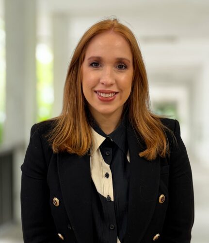 Kathryn Williams, a woman with straight red hair wearing a black blazer and white-collared shirt, stands in a well-lit corridor, smiling at the camera.
