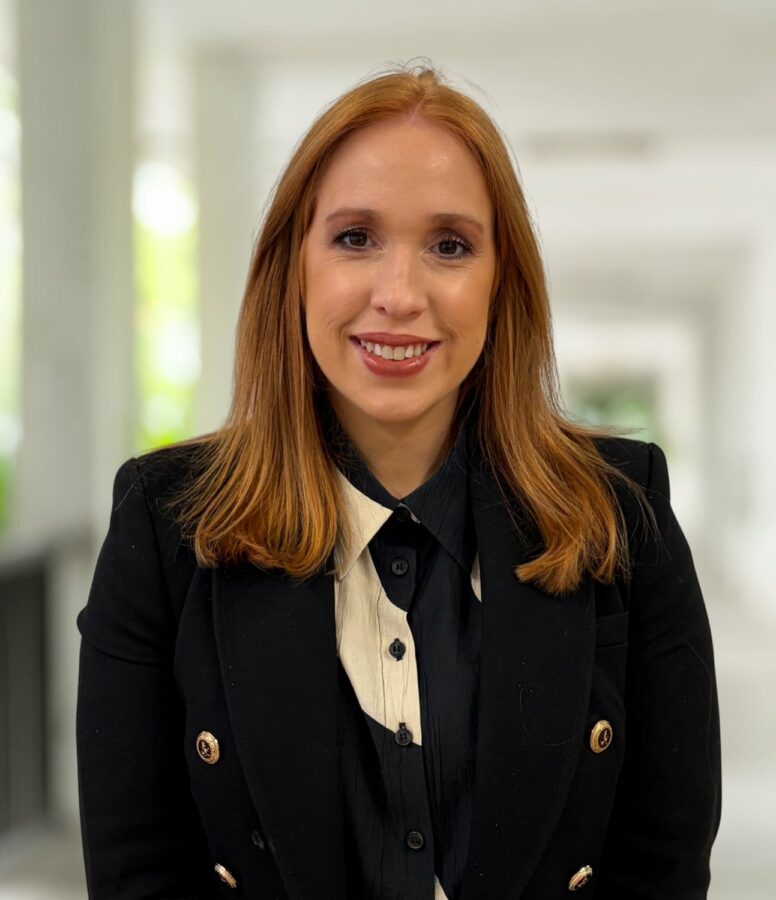 Kathryn Williams, a woman with straight red hair wearing a black blazer and white-collared shirt, stands in a well-lit corridor, smiling at the camera.