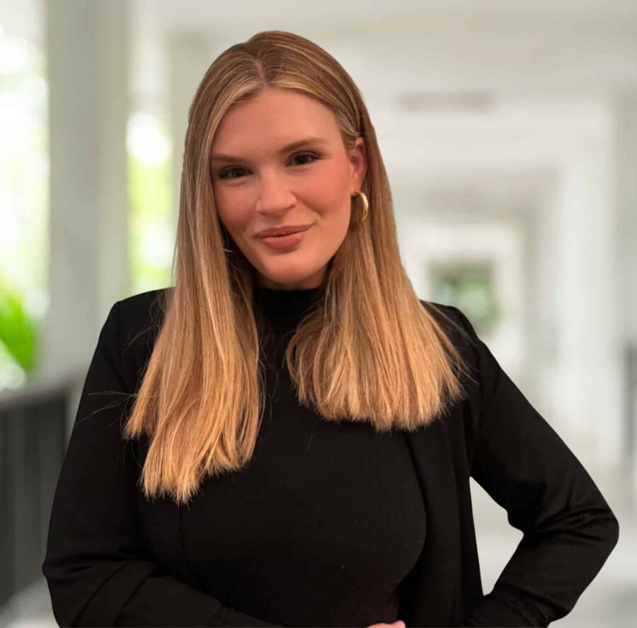 Claire Butcher, with straight blonde hair, wearing a black top and jacket, stands in a bright corridor with a neutral expression.
