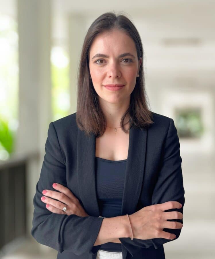 Claudia Lemke, a woman with straight brown hair wearing a black blazer, stands with arms crossed, facing the camera in a well-lit indoor setting with a blurred background.