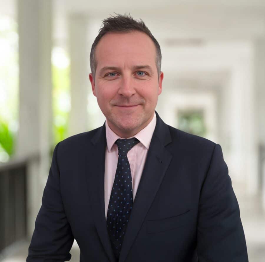 David Howells, in a dark suit and polka dot tie, stands indoors in a bright corridor, facing the camera and smiling slightly.