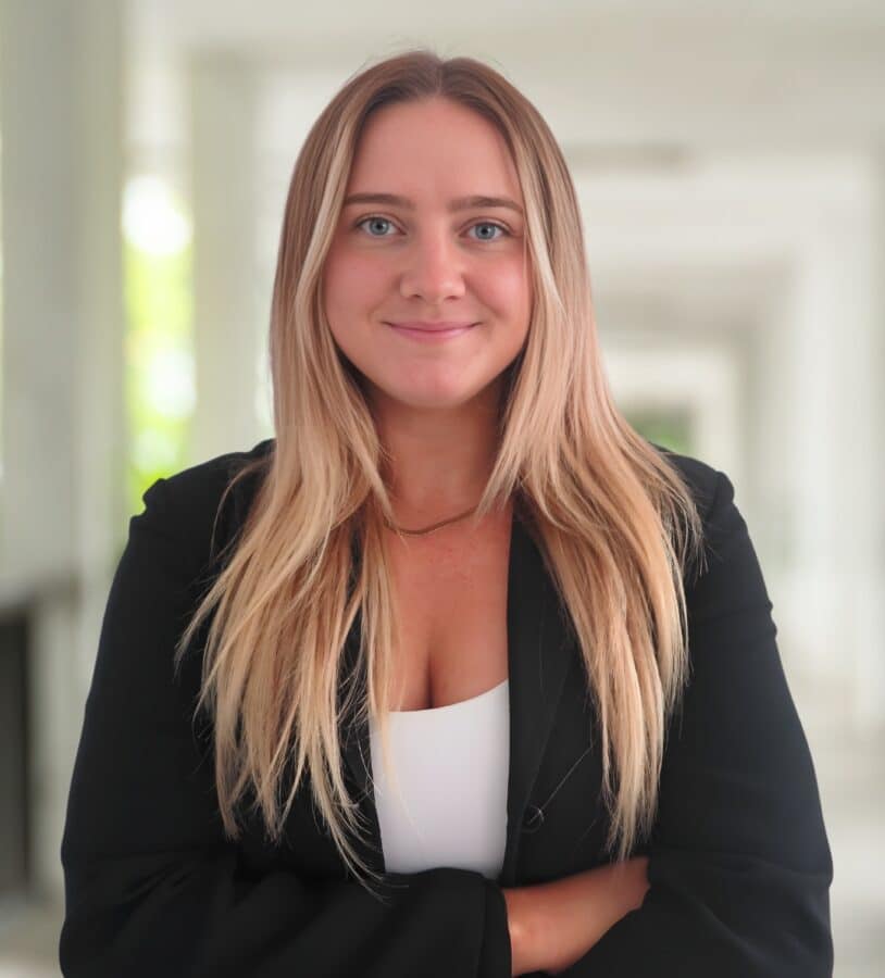 Ellie LaFountain, a woman with long blonde hair, wearing a black blazer and white top, stands indoors with her arms folded, smiling at the camera.