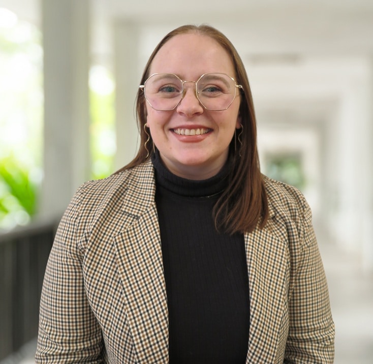 Haley Gibson, with long brown hair, glasses, and a checked blazer over a black polo neck, smiles whilst standing in a bright indoor corridor.