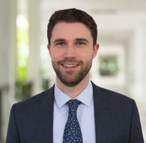 Manuel Preg, in a navy suit and patterned tie, stands indoors, smiling at the camera against a bright, blurred background.