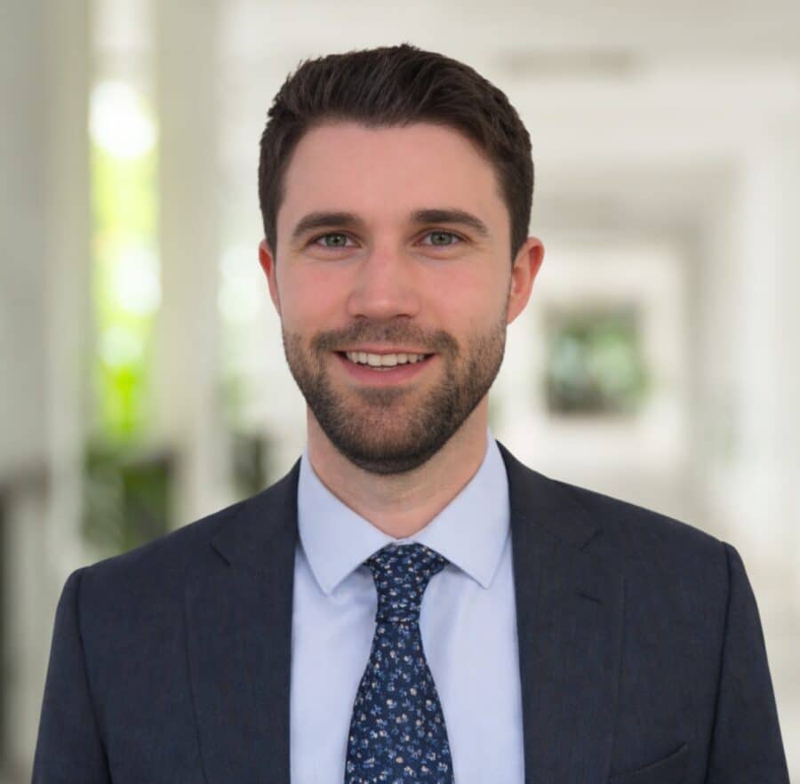 Manuel Preg, in a navy suit and patterned tie, stands indoors, smiling at the camera against a bright, blurred background.
