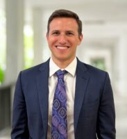Adam Nuzie, dressed in a navy blue suit and patterned tie, stands smiling in a bright corridor with blurred columns and greenery in the background.