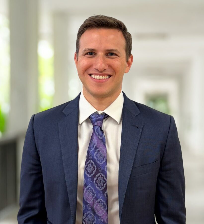 Adam Nuzie, dressed in a navy blue suit and patterned tie, stands smiling in a bright corridor with blurred columns and greenery in the background.