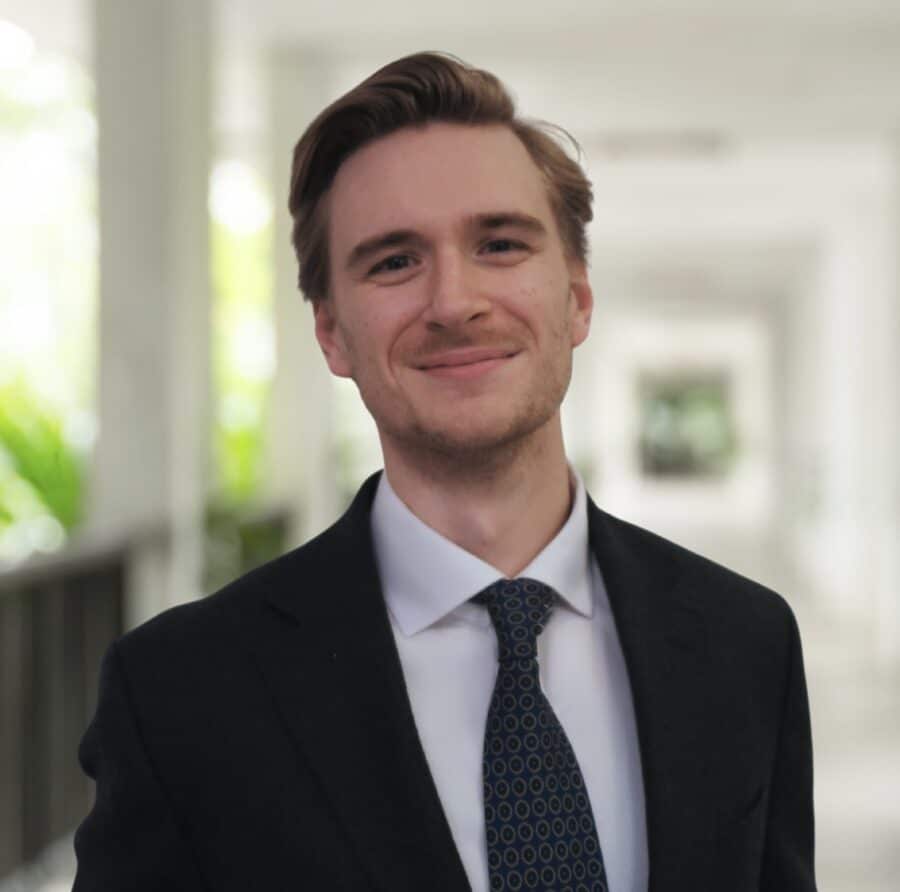 Rupert Haffenden, in a dark suit and patterned tie, stands in a bright corridor with greenery in the background, smiling at the camera.