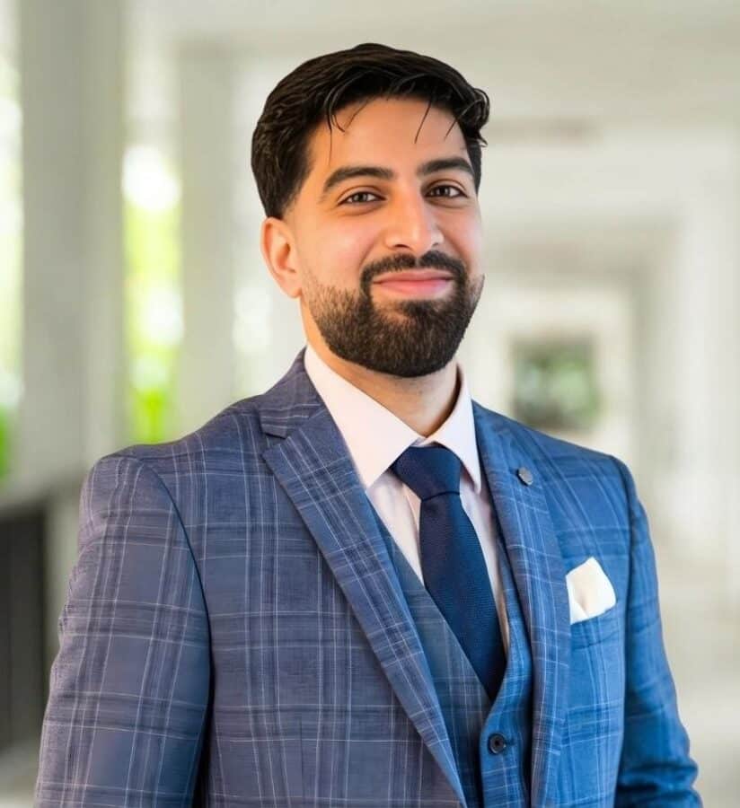 A man with dark hair and a beard, wearing a blue checked suit, waistcoat, and tie, stands indoors with a neutral expression—resembling Sahar Akhtar.