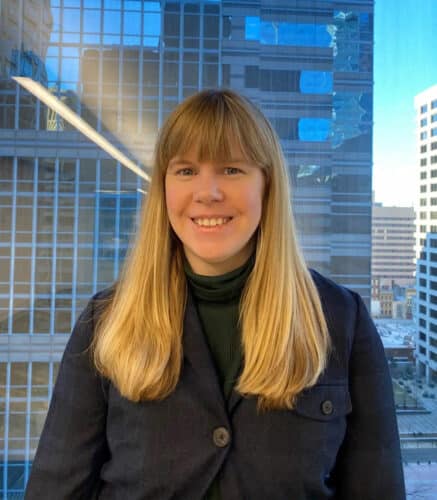A woman with long blonde hair and a dark jacket stands indoors in front of a window with city buildings in the background.