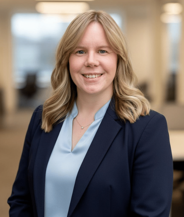 A woman with blonde hair, wearing a light blue blouse and navy blazer, smiles at the camera in an office setting.