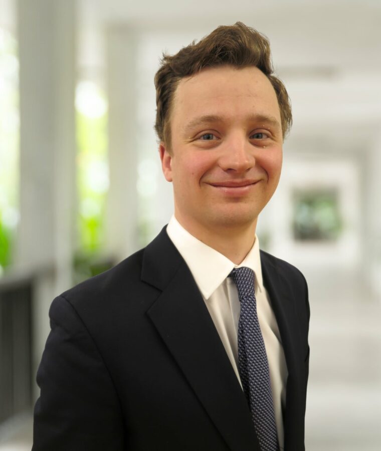 A young man in a dark suit and tie stands in a bright corridor, smiling at the camera.