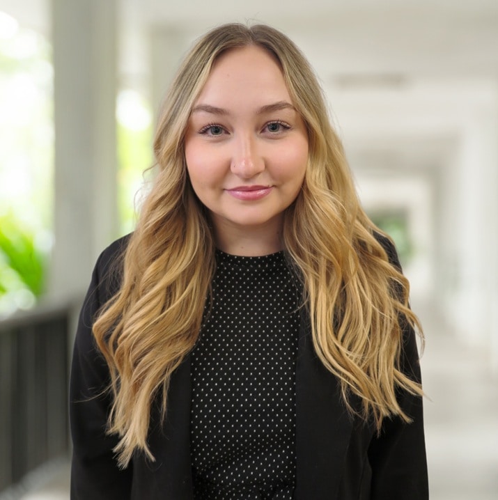 A woman with long blonde hair wearing a black blazer and a black patterned top stands in a bright corridor, looking at the camera.