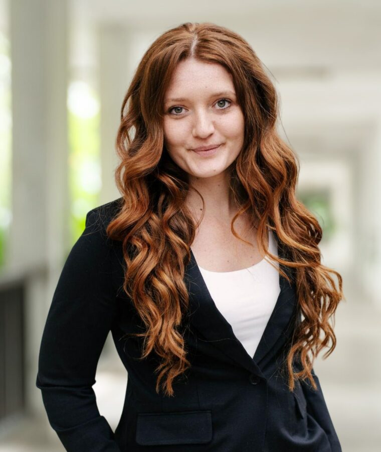 A woman with long wavy red hair, wearing a black blazer and white top, stands in a bright corridor, looking at the camera with a slight smile.