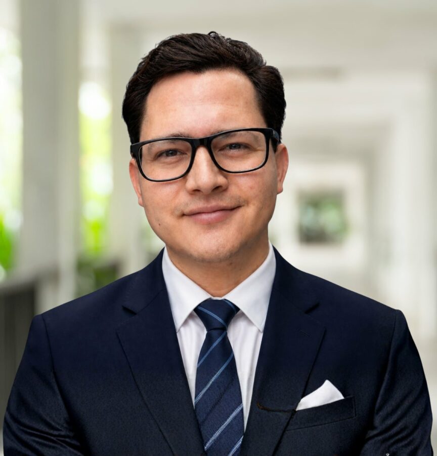Rafael Alvarado, in a dark suit, white shirt, and striped tie, wearing glasses, stands confidently in a bright corridor with a blurred background.