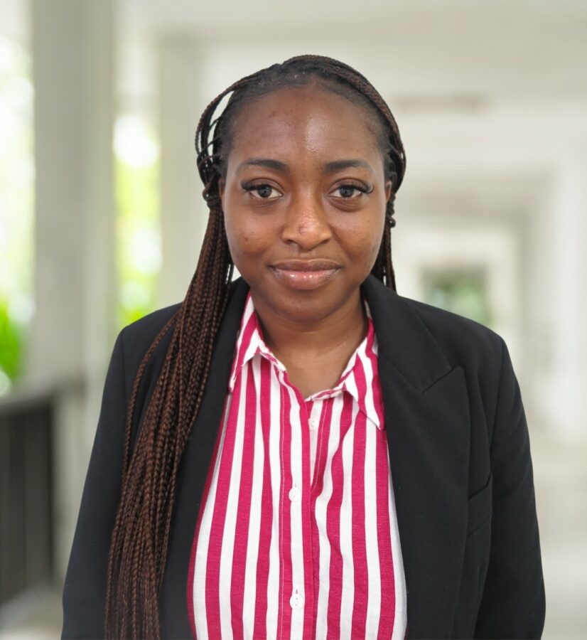 Feyisayo Adetayo, a woman with long plaits, stands in a corridor wearing a black blazer over a red and white striped shirt, looking confidently at the camera.