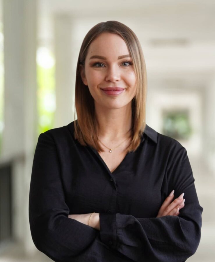 Woman with straight, shoulder-length hair and a black shirt, stands indoors with her arms folded, smiling slightly. The background is blurred.