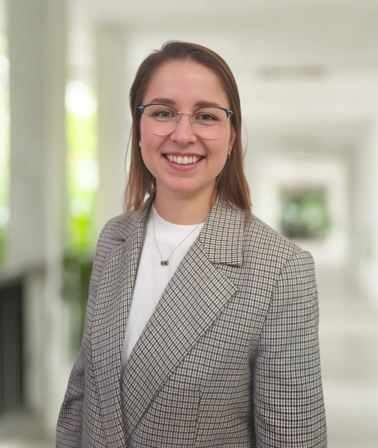 A woman wearing glasses and a checked blazer smiles whilst standing in a bright corridor with blurred background.