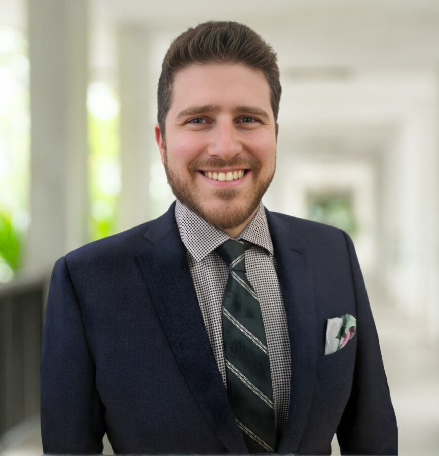 A man in a dark suit, checked shirt, and striped tie stands smiling in a bright corridor with blurred columns and greenery in the background.