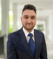 A man in a navy blue suit and tie stands in a bright, covered outdoor corridor, looking at the camera with a neutral expression.