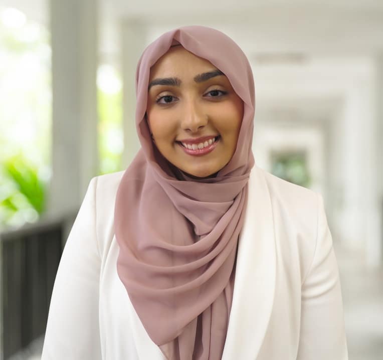 A woman wearing a mauve hijab and white blazer smiles whilst standing in a bright, corridor-like space with greenery in the background.