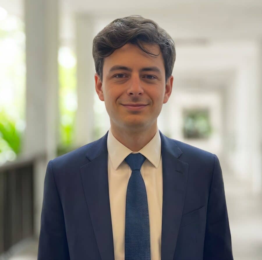 A man in a blue suit and tie stands in a bright, corridor-like setting with greenery visible in the background.