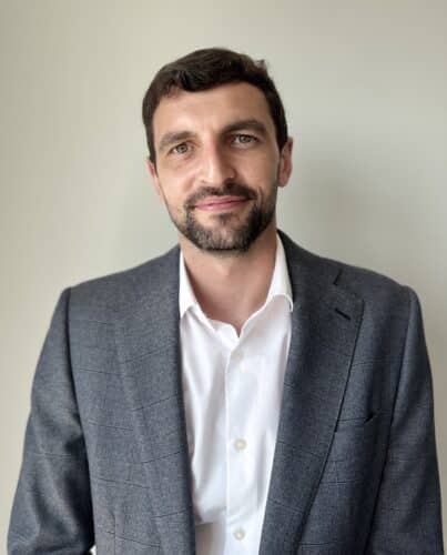 Man with short dark hair and beard wearing a grey suit jacket over a white shirt, standing in front of a plain light-coloured background.
