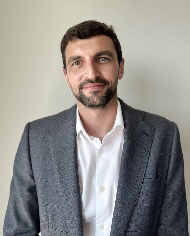Man with short dark hair and beard wearing a grey suit jacket over a white shirt, standing in front of a plain light-coloured background.