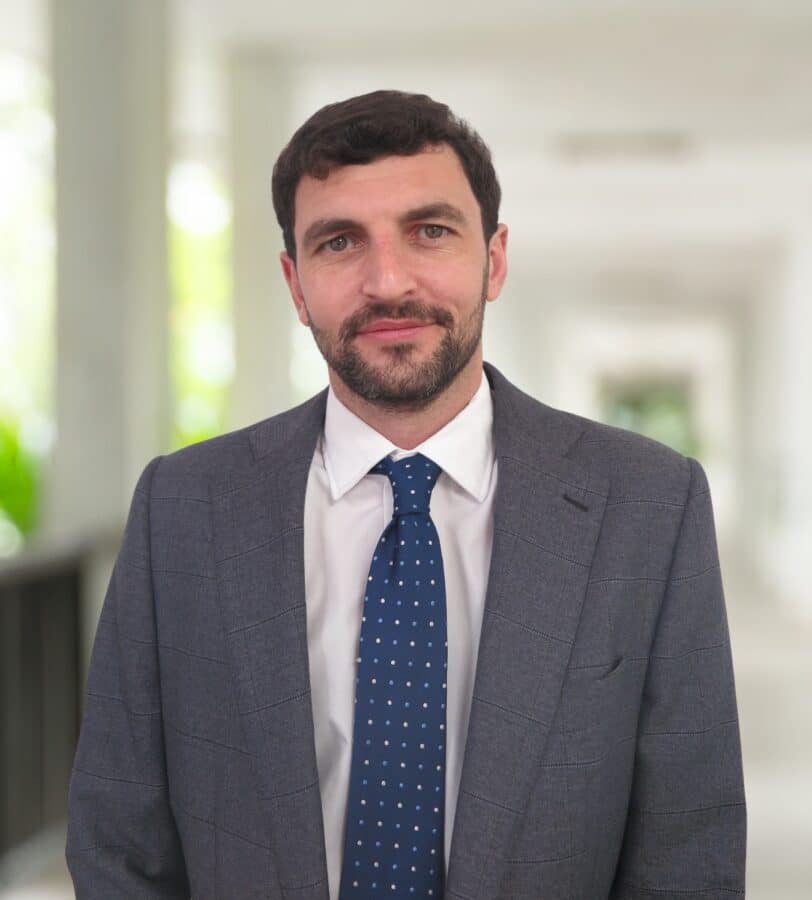 Man with short dark hair and beard wearing a grey suit, white shirt, and blue polka dot tie, standing in a bright, blurred corridor.