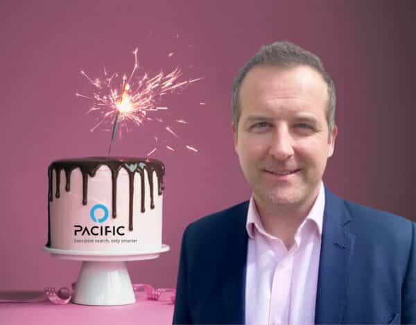 A man in a suit stands in front of a pink cake with chocolate drip, a lit sparkler, and the Pacific company logo, against a pink background.