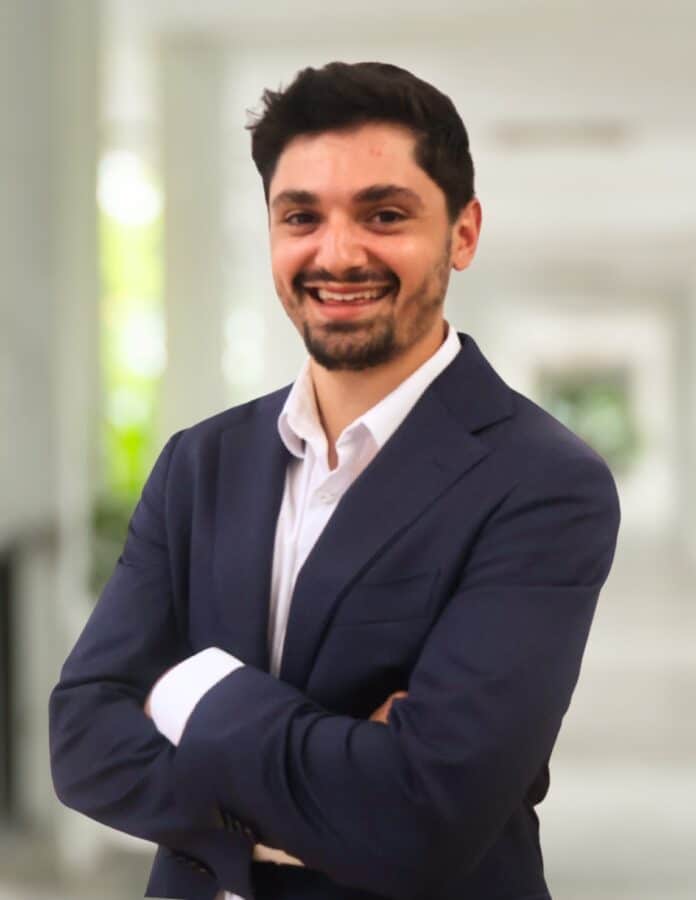 A man in a navy suit and white shirt stands indoors with arms folded, smiling at the camera. The background is softly blurred.