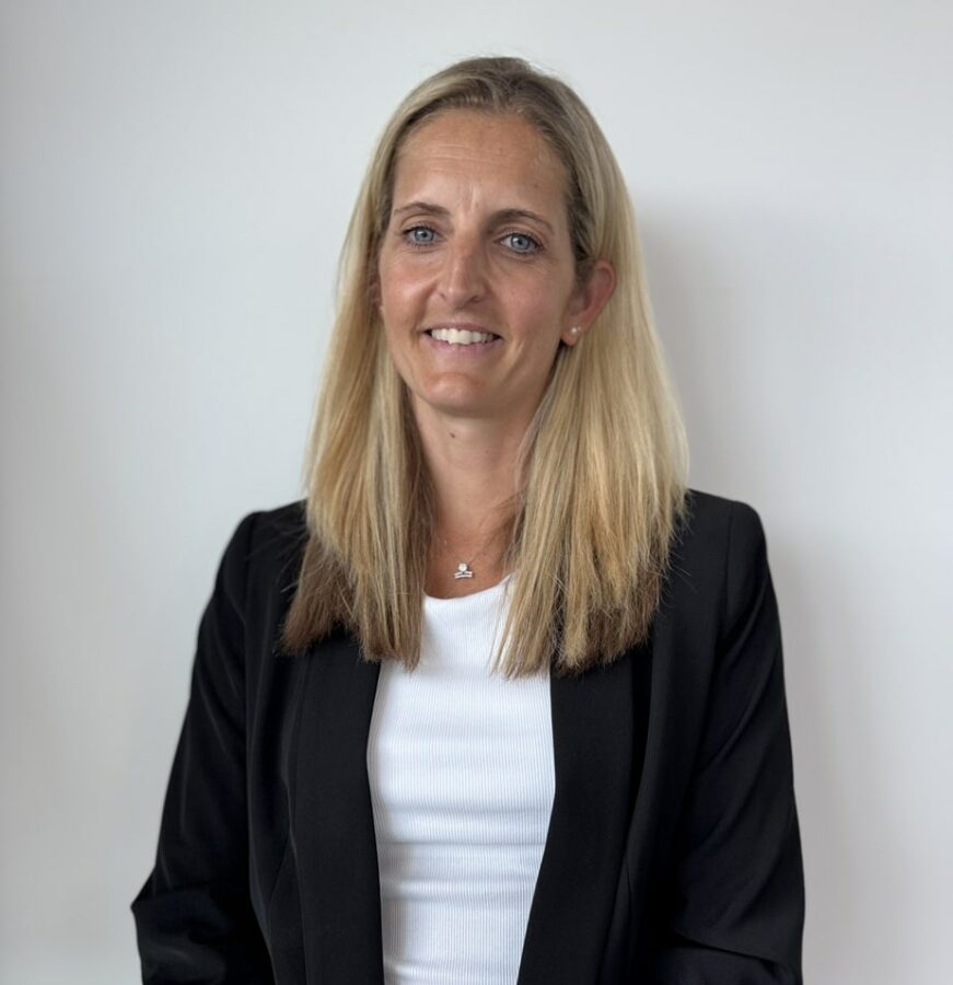 A woman with long blonde hair, wearing a black blazer over a white top, stands in front of a plain white background, smiling at the camera.