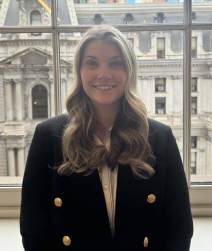 A woman with long wavy hair in a black blazer stands indoors in front of a window, with a historic stone building visible in the background.