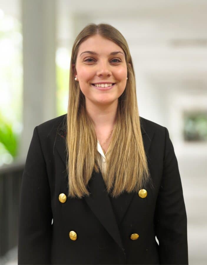 A woman with straight blonde hair, wearing a black blazer with gold buttons, stands indoors in a well-lit corridor, smiling at the camera.