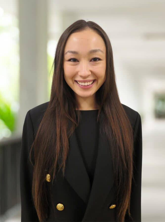 Woman with long brown hair wearing a black blazer stands indoors, smiling at the camera with a blurred background.