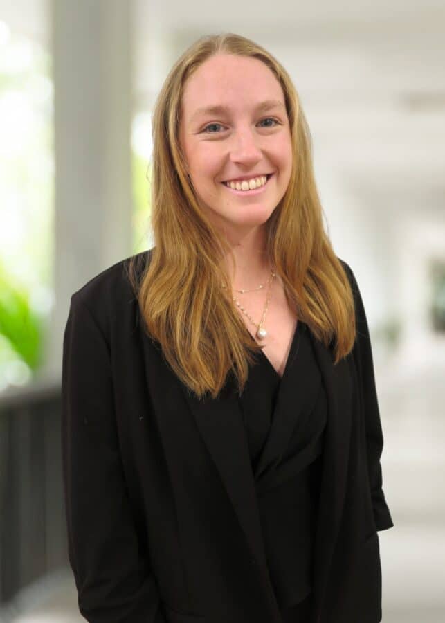 A woman with long, light brown hair wearing a black blazer and necklace stands indoors, smiling at the camera. The background is softly blurred.