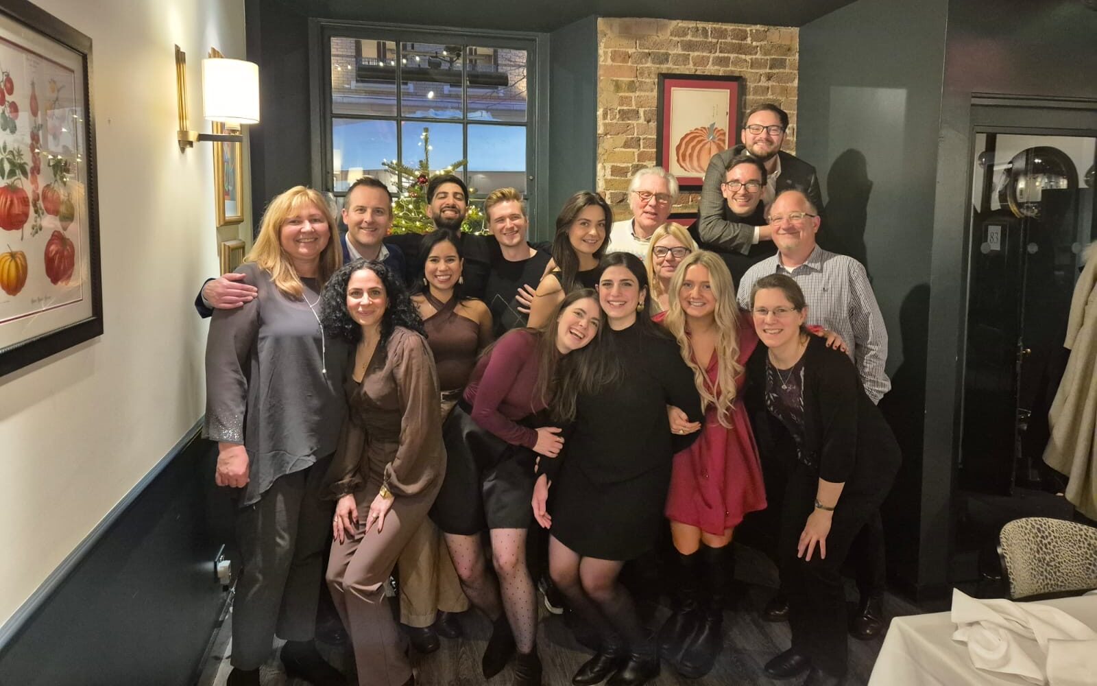 A group of fifteen people posing together and smiling inside a restaurant, with festive decorations and framed artwork on the walls—come join our dynamic team!.