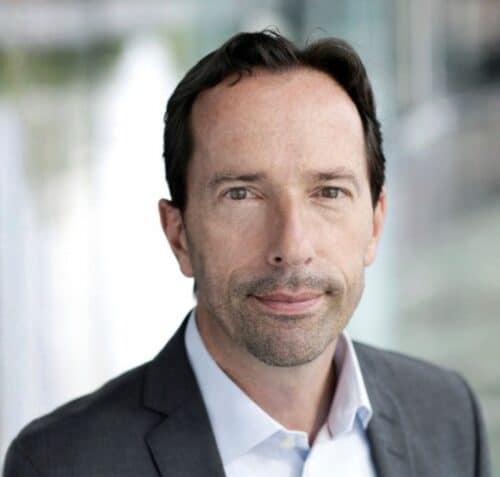 A man with short dark hair and light stubble, wearing a grey suit jacket and white shirt, stands in front of a blurred indoor background.