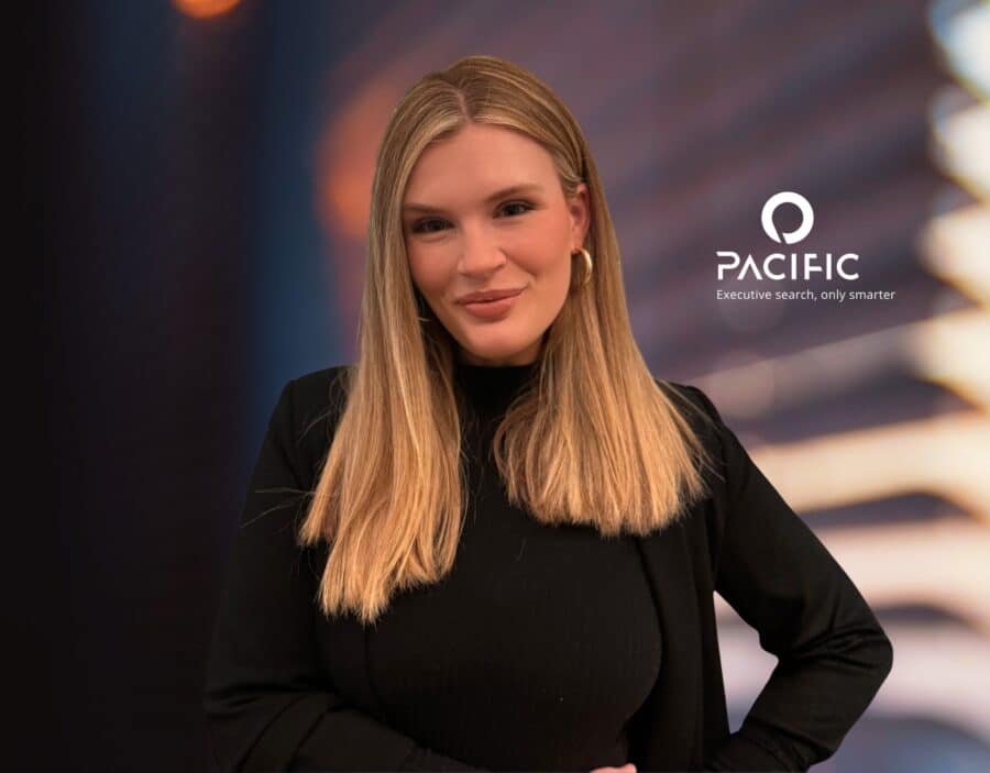 A woman with long blonde hair in a black outfit stands in front of a blurred background with the Pacific company logo and the tagline "Executive search, only smarter" on the right.