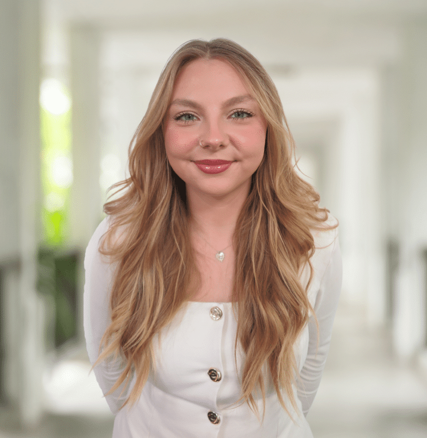 A young woman with long blonde hair, wearing a white top with buttons, stands indoors in a bright corridor, smiling at the camera.