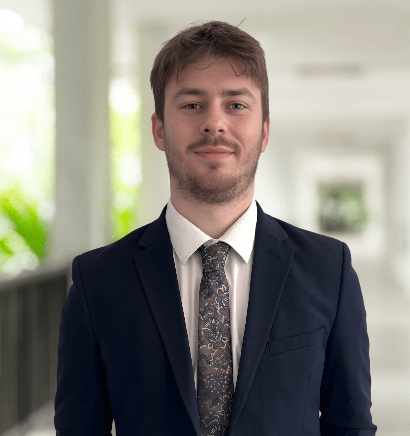 A young man in a dark suit, white shirt, and patterned tie stands in a bright corridor with blurred background and greenery visible.