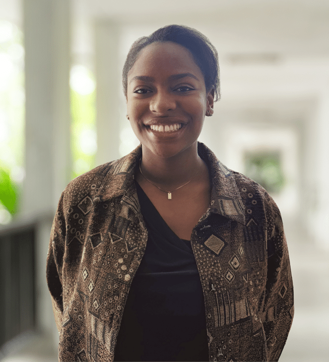 A woman stands in a corridor, smiling at the camera. She is wearing a patterned brown jacket over a black top. The background is softly blurred.