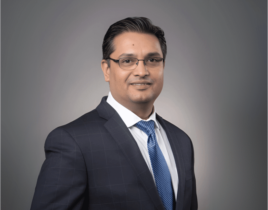 Sameer Pagnis, wearing glasses, a dark checked suit, white shirt, and blue tie, poses for a professional studio portrait against a grey background.