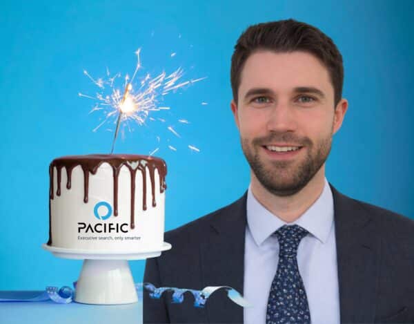 A man in a suit stands next to a chocolate-drizzled cake with a sparkler on top and the Pacific logo, celebrating a decade of growth against a blue background.