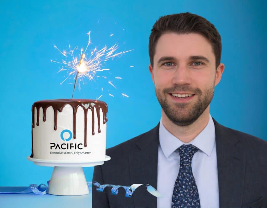 A man in a suit stands next to a chocolate-drizzled cake with a sparkler on top and the Pacific logo, celebrating a decade of growth against a blue background.