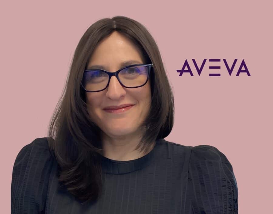 A woman with long brown hair and glasses smiles in front of a pink background with the AVEVA logo.