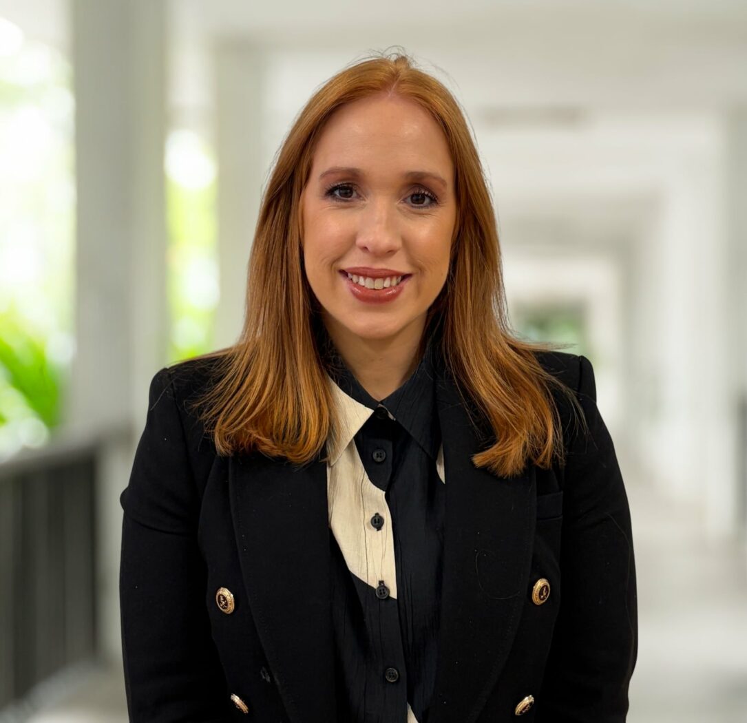 A woman with straight red hair, wearing a black blazer and white shirt, stands in a bright corridor with blurred background, ready to welcome you to join our dynamic team.