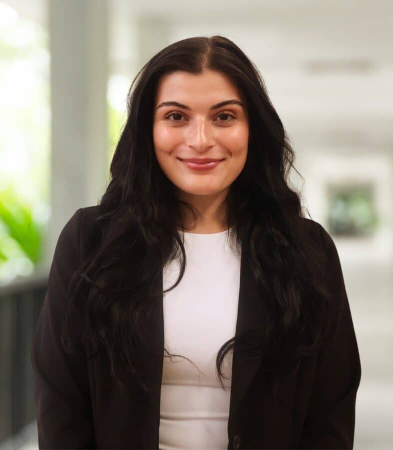 A person with long dark hair, wearing a black blazer and white top, stands indoors in a bright, blurred corridor.