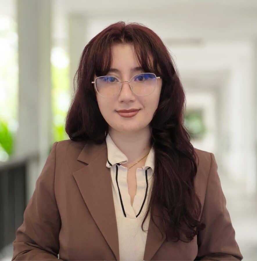 A person with long brown hair and glasses, wearing a brown blazer and a light-coloured blouse, stands in a bright indoor corridor.