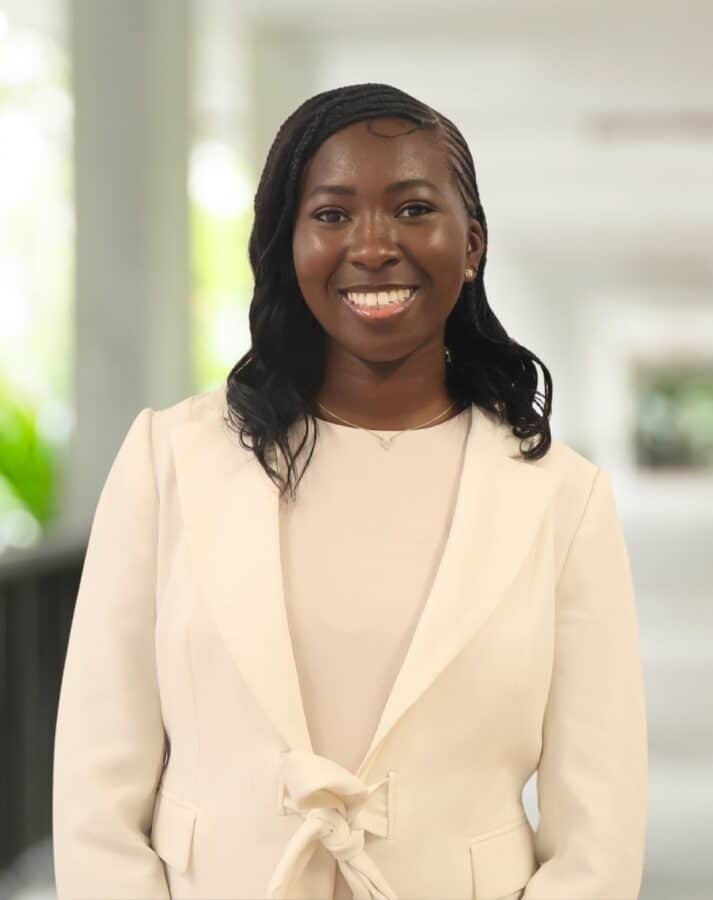 A woman with long black hair wearing a cream-coloured suit jacket and light top stands in a bright corridor, smiling at the camera.